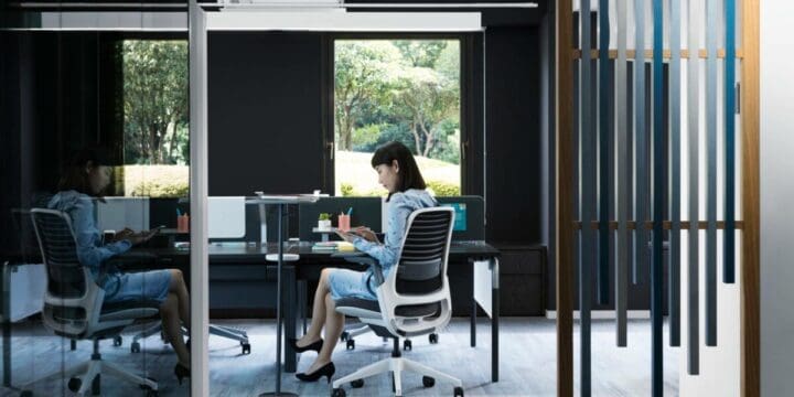 A modern office space with glass partitions where a woman is seated at a desk, working intently, with a reflection of her on the adjacent glass and a view of green trees outside.