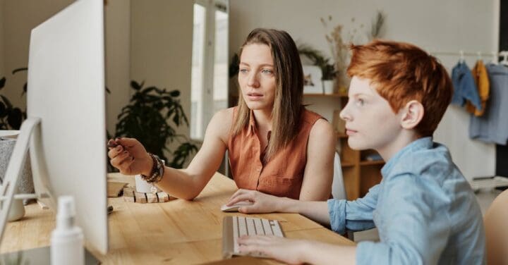 features two individuals in an indoor setting.
The woman in the foreground has long brown hair and is wearing a rust-colored sleeveless top. She appears focused, looking slightly downwards, possibly at a computer screen or a document. Beside her is a young boy with bright orange-red hair, wearing a blue shirt. He is looking off to the side, seemingly attentive to something or someone out of the frame. The setting appears to be a workspace or home office, given the presence of a computer monitor and other desk accessories. There are also plants and a room divider in the background, and a few items of clothing are hanging up, suggesting this might be a multifunctional space.
The lighting is natural, and the overall ambiance of the image gives a sense of concentration and calm.