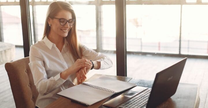 a woman sitting at a desk in a spacious room with large windows. The ambient lighting suggests it might be near sunset. She's wearing glasses and a white shirt, smiling as she looks at something in her hand, perhaps a pen or a small object. In front of her is an open notebook, and a laptop is also present on the desk. enhance personal productivity.