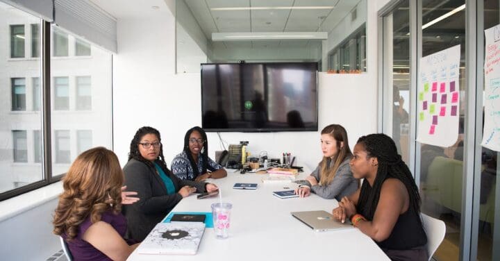 a group of women seated around a conference table in what appears to be a modern office space. The room is brightly lit with large windows, and there is a television screen on one wall. The table has various office supplies, including laptops, notebooks, and pens. One of the walls features a whiteboard with colorful post-it notes and writing on it, indicating a brainstorming or planning session. The women seem to be engaged in a discussion. Enhance personal productivity